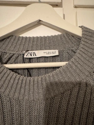 Grå croppad stickad tröja från Zara - Säljer en grå croppad stickad tröja från Zara med ribbad struktur och rund hals. Tröjan har långa ärmar och en lös passform som ger en chill vibe. Perfekt att matcha med höga jeans eller kjol för en snygg look. Använd fåtal gånger.