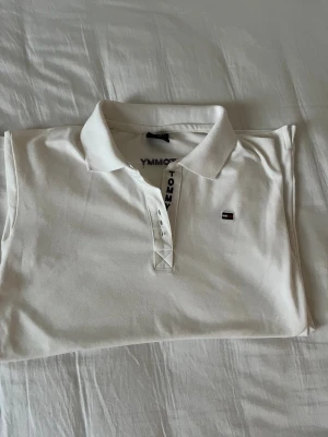 Vit pikétröja Tommy Hilfiger - XXL i barnstorlek! Snygg vit pikétröja från Tommy Hilfiger med klassisk krage och knappar framtill. Broderad logga på bröstet och TOMMY-text längs knappslån. Mjuk bomullskvalitet som är skön mot huden. Perfekt för dig som gillar stilrena och tidlösa plagg.