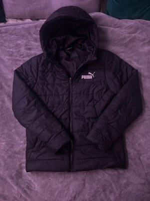 Svart quiltad pufferjacka från Puma - Svart pufferjacka från Puma med huva och snyggt quiltad design. Jackan har dragkedja framtill och Puma-logga på både bröstet och ryggen. Tillverkad i lätt polyester som håller dig varm och bekväm under kyliga dagar.