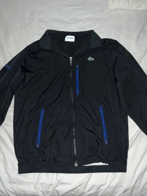 Svart vindjacka från Lacoste Sport - Svart vindjacka från Lacoste Sport med blå detaljer på dragkedjorna och logga på ärmen. Klassisk krage, tre fickor med dragkedja och ikoniska krokodilloggan på bröstet. Perfekt för dig som gillar sportig och clean stil.
