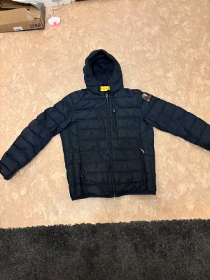 Mörkblå dunjacka Parajumpers med huva - Snygg mörkblå dunjacka från Parajumpers med huva och coolt patch på ärmen. Jackan har quiltad design, dragkedja framtill och två sidofickor med dragkedja. Insidan är grå och märkt med 'Super Lightweight'. Perfekt för kalla dagar.        Vill säga bara att säljer den billigt eftersom den är L i Barnstorlek så för 14 15 åringar den har jag växt ut små skråmor men annars bra skicka går att skanna helt äkta kan fråga frågor i dm. Pris kan diskuteras lite. Snabb frakt skickar direkt elelr dagen efter