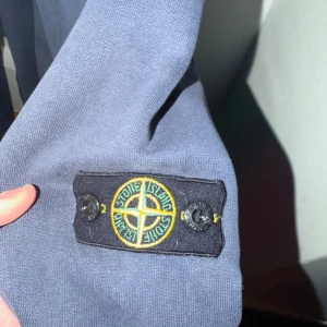 Mörkblå hoodie från Stone Island - Säljer en mörkblå hoodie från Stone Island med klassisk patch på ärmen. Tröjan har huva, dragkedja framtill och två fickor. Materialet är mjuk bomull och passformen är normal. Perfekt för dig som gillar streetwear och snygga detaljer. Strl är mellan 12-15 år. Kommer stå strl 50 för jag inte har koll vad det blir.