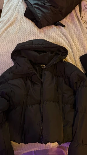 Svart croppad pufferjacka från H&M - Svart pufferjacka från H&M med croppad passform och huva. Jackan har dragkedja framtill och snörning vid nederkanten för att justera stilen. Perfekt för kalla dagar och har en modern, loose siluett.