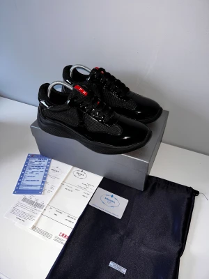 Prada cups helt svarta - Säljer ett par svarta Prada cups med glansiga detaljer och meshpaneler. Skorna har en chunky sula, snörning och röda logodetaljer på plös och sula. Materialmix av skinn och syntet ger en lyxig och sportig vibe. Perfekta för dig som vill sticka ut med exklusiva sneakers.