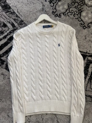 Vit kabelstickad tröja Polo Ralph Lauren - Snygg vit kabelstickad tröja från Polo Ralph Lauren med klassisk rund hals och broderad marinblå logga på bröstet. Tröjan har ribbade muddar och är långärmad, perfekt för dig som gillar stilrena och tidlösa plagg.