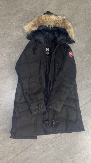 Svart dunjacka från Canada Goose - Säljer en svart dunjacka från Canada Goose med huva och snygg pälsdetalj. Jackan har lång passform, dragkedja och knappar framtill samt logotyp på ärmen. Perfekt för kalla dagar och riktigt stilren look.