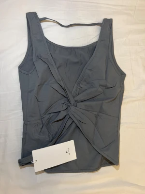 Grå linnetopp med twist från La Mania - Snygg grå linnetopp från La Mania med twistad detalj framtill som ger en cool look. Toppen är ärmlös och har en rak, något loose passform. Materialet känns mjukt och lätt, perfekt för varmare dagar. Enkel design med breda axelband och diskret logga på etiketten.