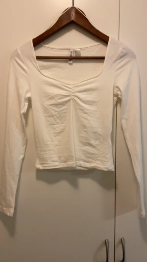 Vit långärmad topp från Divided - Säljer en vit långärmad topp från Divided med snygg v-ringning och rynkad detalj framtill. Modellen är croppad och har en mjuk, skön känsla i bomullsmaterial. Perfekt för dig som gillar enkel och stilren look med lite edge.