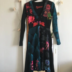 Svart mönstrad Desigual klänning - Unik svart klänning från Desigual med färgstarka blommor, zebramönster och cykeltryck. V-ringad, långärmad och med coola blå detaljer på ena ärmen. Materialet är mjukt och stretchigt, perfekt för dig som gillar statement-plagg med kreativ design.