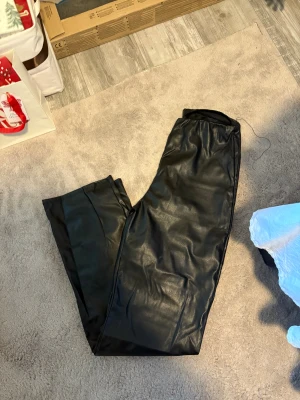 Svarta byxor i läderimitation - Säljer ett par svarta byxor i läderimitation med resår i midjan. Byxorna har en rak och loose passform, perfekta för dig som vill ha en edgy och trendig look. Materialet är syntetiskt och ger en cool läderkänsla.