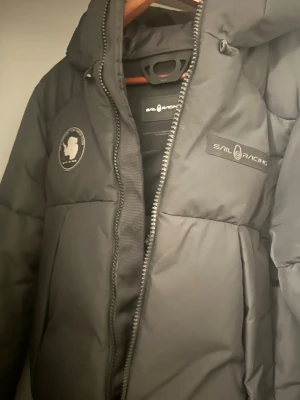 Svart pufferjacka från Sail Racing - Svart pufferjacka från Sail Racing med huva och snygga logopatchar på bröstet och ärmen. Jackan har dragkedja framtill och är tillverkad i polyester. Perfekt för kalla dagar med sin tjocka och quiltade design. Stilsäker look med clean finish och diskreta detaljer.