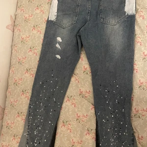 Gallery Dept. jeans  - Unika blå jeans från Gallery Dept. med coola vita och svarta färgstänk, slitna detaljer och kontrasterande mörka paneler längs benen. Bakfickorna har vitmålade partier för extra edge. Perfekta för dig som vill sticka ut med din stil.