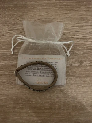 Tennarmband från Arctic Collection - Unikt tennarmband från Norrbotten, handsytt på mjukt brunt renskinn med flätad tenntråd och silverpärlor. Stängs med knapp av horn. 