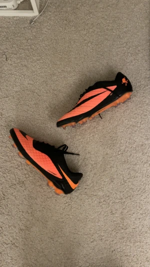Nike Hypervenom Phantom fotbollsskor - Säljer ett par Nike Hypervenom Phantom fotbollsskor i svart och orange med coola döskalle-detaljer på hälen. Skorna har snörning och platt sula med dobbar för gräsplan. Ovandelen är tillverkad i syntetmaterial med textur för bättre bollkontroll.