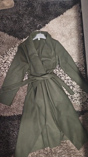 Mörkgrön kappa med bälte från New Collection -  mörkgrön kappa från New Collection med lång krage och bälte i midjan. Jackan har långärmade ärmar.Perfekt för dig som gillar stilrena och enkla plagg med lite edge. Helt ny, aldrig använd. Köpt egentligen för 800kr. Pris kan diskuteras