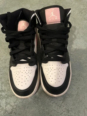 Nike Air Jordan 1 svart/vit/rosa - Nike Air Jordan 1 sneakers i svart, vit och ljusrosa. Klassisk high-top modell med rosa swoosh och detaljer på tungan. Skorna har snörning, perforerad tå och platt sula. Materialet är skinn och syntet, vilket ger en snygg och sportig vibe.