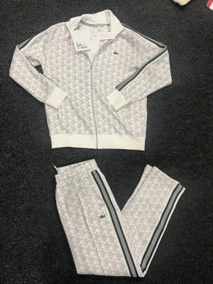 Vita mönstrade mjukisbyxor från Lacoste - Snygga vita mjukisbyxor från Lacoste med geometriskt mönster och gröna samt grå ränder längs sidorna. Byxorna har resår och snörning i midjan samt klassisk Lacoste-logga på benet. Perfekta för en sportig och chill look.finns Alla storlekar 