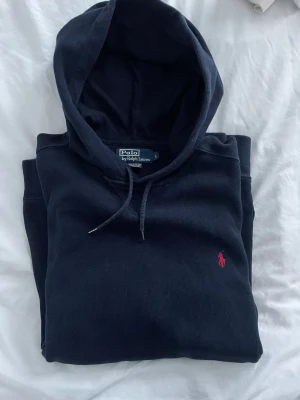 Mörkblå hoodie från Polo Ralph Lauren - Säljer en mörkblå hoodie från Polo Ralph Lauren med klassisk röd logobrodering på bröstet. Tröjan har huva med snörning, stor magficka och långa ärmar. Perfekt för chill dagar och enkel att matcha med jeans eller joggers.