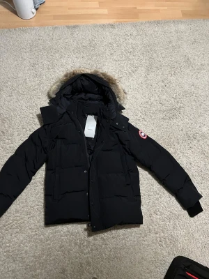 Svart Wyndham Parka från Canada Goose - Säljer en svart Wyndham Parka från Canada Goose med pälskantad huva och klassisk logga på ärmen. Jackan är dunfodrad, har dragkedja och knappar framtill samt flera fickor. Perfekt för kalla vinterdagar och har en riktigt clean look.