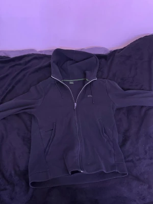 Svart hoodie från BOSS med dragkedja - Svart hoodie från BOSS med hel dragkedja framtill och två fickor. Tröjan har huva med snörning och långa ärmar. Materialet är mjukt och känns som bomull, perfekt för chill dagar. Diskret BOSS-logga på bröstet och stilren design.
