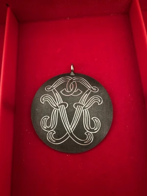 Runt halsband från Svenskt Tenn - Unikt runt halsband i tenn från Svenskt Tenn, handarbete med snyggt graverat monogram på framsidan. Hängsmycket har en cool vintagekänsla och är i silverfärgad ton. Perfekt för dig som gillar klassisk design med en twist.