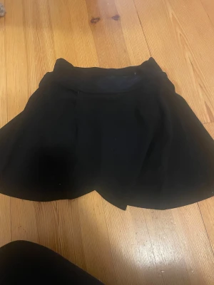 Svart kortkjol - Svart, enkel och stilren kortkjol från SHEIN i storlek XS. Kjolen har en lätt utsvängd form och är tillverkad i mjukt syntetmaterial. Perfekt för dig som gillar minimalistisk och clean look. Passar dig som vill ha en klassisk svart kjol med skön passform. Shorts under kjolen.