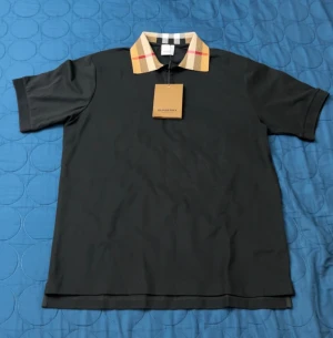 Svart Burberry skjorta med rutig krage - Säljer en svart pike från Burberry med klassisk rutig krage i beige, svart, vitt och rött. Piken har korta ärmar och en stilren, rak passform. Materialet är mjukt och andas, perfekt för en clean och trendig look. Helt ny oanvänd. 