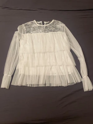 Vit blus med spets och pärlor💕 - Superfin vit blus i transparent mesh med långa ärmar, volanger och små pärlor. Överdelen har broderade blommor i spets och knytning bak i nacken. Perfekt för dig som gillar romantisk och drömmig stil. Säljes till bästa pris! Liknar Ida Sjöstedt blusen. Se sista bild för den lilla defekten men syns knappt när man har på sig den då volangerna täcker det💕