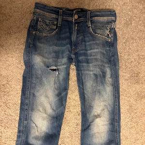 Blå Replay anbass jeans med slitning - Säljer ett par blå jeans från Replay med snygg slitning och hål på höger lår. Klassisk femficksmodell, normal passform och raka ben. Jeansen har ljusa tvättade partier och tydliga sömmar. Perfekta för en avslappnad streetstil.