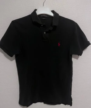 Svart Polo Ralph Lauren pike S - Svart pike från Polo Ralph Lauren i slim fit . Pike. är gjord i 100% mjuk bomull. På bröstet finns den ikoniska röda broderade Ralph Lauren loggan.