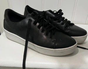 Svarta sneakers från XIT, storlek 39 - Svarta sneakers från XIT med stilren design och vita, platta sulor. Skorna har snörning och är gjorda i ett slätt material som liknar skinn. Perfekta för dig som gillar en clean och klassisk look.