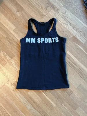 Svart linnetopp från MM Sports - Svart linne från MM Sports med vit texttryck både fram och bak. Modellen har brottarrygg och är figurnära. Materialet känns mjukt och stretchigt..