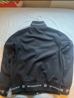 Svart bomberjacka Givenchy S - Givenchy bomberjacka i svart med röda och vita ränder längs ärmarna. Jackan har dragkedja framtill, ribbade muddar och Givenchy-logga på krage och nederkant. Snygg, sportig look med normal passform och lång ärm. Perfekt för dig som gillar streetwear och designer.