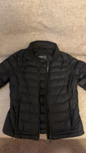 Svart pufferjacka från Sail Racing - Svart jacka från Sail Racing med quiltad design och dragkedja framtill. Jackan har en stilren, kortare passform och ribbad krage. Materialet är syntet och jackan har två sidofickor med dragkedja. Perfekt för dig som gillar en clean och sportig look.