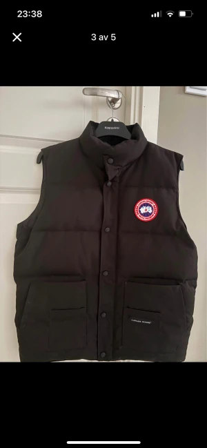Svart dunväst från Canada Goose - Svart dunväst från Canada Goose med klassisk logga på bröstet. Västen har hög krage, två stora fickor framtill och stängs med knappar. Perfekt för lager-på-lager och håller dig varm under kyliga dagar.
