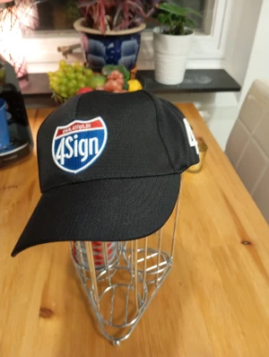 Svart keps från 4Sign - Svart keps med böjd skärm och broderad 4Sign-logga i blått, vitt och rött framtill. Klassisk form med justerbar passform bak. Perfekt för dig som vill ha en enkel och stilren keps med tydlig branding.