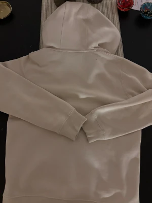 Beige hoodie från Sail Racing - Snygg beige hoodie från Sail Racing med hel dragkedja och huva. Tröjan har svarta detaljer vid dragkedjan och logga på bröstet. Mjukt material och praktiska fickor framtill, perfekt för chill dagar. Passar dig som gillar stilren och sportig look.