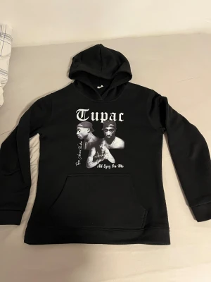 Svart Tupac hoodie med tryck - Svart hoodie med stor vit Tupac-tryck på bröstet och motiv från albumet 'All Eyez On Me'. Baksidan har låtlista i vitt tryck. Klassisk huva och magficka, långärmad och loose passform. Perfekt för dig som gillar streetwear och hiphop.