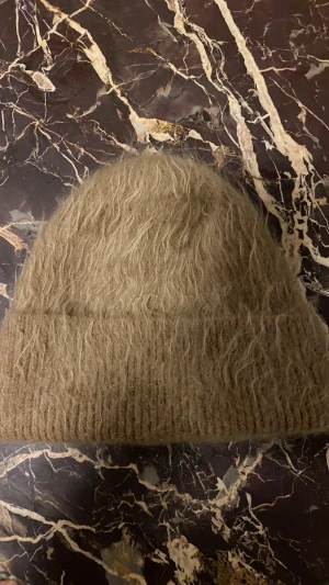 Beige fluffig stickad mössa - Säljer en beige, fluffig och mjuk mössa med ribbad kant. Mössan har en enkel och stilren design med en lite lurvig yta som ger en cool och trendig look. Perfekt för kalla dagar när du vill hålla dig varm och snygg. Märke -ARKET; st. One siden; 45% Mocher. 