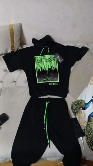 Svart croppad hoodie från Guess - Svart croppad hoodie från Guess med korta ärmar och huva. Framsidan har ett stort grönt tryck med logga och coolt grafiskt mönster. Neongröna snören i huvan ger extra edge. Perfekt för dig som gillar streetstyle och vill sticka ut.