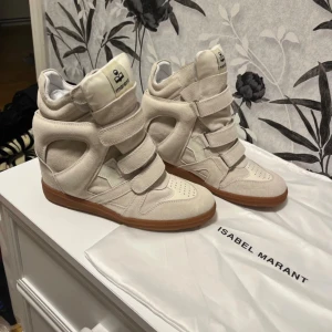 Beige sneakers med kilklack Isabel Marant - Snygga beige sneakers från Isabel Marant med kilklack och platt gummisula. Skorna har flera breda kardborreband, rund tå och är tillverkade i mocka med detaljer i syntet. Perfekt för dig som vill ha en cool och unik look med extra höjd. Skriv vilken storlek ni har.