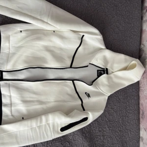 Vit hoodie från Nike - Säljer en stilren vit hoodie från Nike med svarta detaljer och dragkedja. Perfekt för en sportig look. Den har en justerbar huva och två fickor framtill.