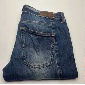 Blå Replay jeans Radula W27 L30 - Snygga blå jeans från Replay, modell Radula. Klassisk femficksdesign med kontrastsömmar och diskreta slitningar. Jeansen har röda detaljer vid bakfickan och är tillverkade i mjukt denimtyg för en bekväm passform.drag kedja på baksidan av byxorna.