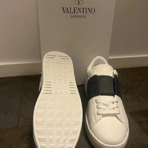 Valentino open - Snygga Valentino Garavani Open sneakers i vitt skinn med bred svart läderdetalj över vristen. Klassisk rund tå, vita snören och ikoniska nitar baktill. Kommer med originalkartong, dustbag och certifikat. Perfekta för dig som vill ha en clean och lyxig look.