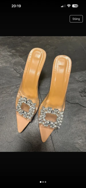 Beige klackpumps med strassdetalj - Säljer ett par beige pumps med spetsig tå och transparent rem över foten, dekorerad med stora glittriga strasstenar. Klacken är hög och gjord i klar plast med stjärnformad design. Perfekt för dig som vill sticka ut med något extra.