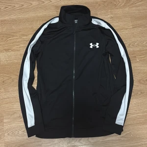 Svart träningsjacka Under Armour - Sportig svart träningsjacka från Under Armour med vita ränder längs ärmarna och logga på bröstet. Den har storlek S. Hör av dig vid intresse!