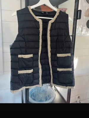 Svart dunväst med beige detaljer - Stilren svart dunväst med beige fransiga kantband längs kanter och fickor. Västen har fyra framfickor och quiltad design som ger en modern look. Perfekt för lager-på-lager och enkel att matcha med olika outfits.