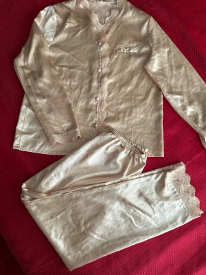 Beige satinpyjamas med spetsdetaljer XL - Snygg tvådelad pyjamas i beige satin med långärmad överdel och byxa. Båda delarna har fina spetsdetaljer vid ärmslut, benslut och längs knäppningen. Överdelen har små pärlknappar och en bröstficka. Perfekt för mysiga kvällar hemma.