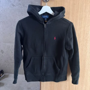 Svart hoodie från Polo Ralph Lauren - Svart hoodie från Polo Ralph Lauren med dragkedja och huva. Klassisk design med rött broderat Polo-logo på bröstet och två fickor framtill. Tillverkad i mjuk bomull. På bilderna ser tröjan mer blekt/urtvättad ut än vad den är i verkligheten. 