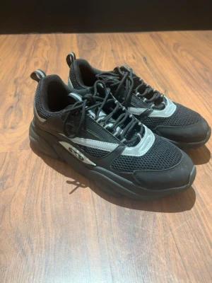 Dior b22 - Snygga svarta sneakers med chunky sula och silverfärgade detaljer. Ovandelen är i mesh och syntetmaterial för bra andningsförmåga och komfort. Skorna har svarta skosnören och en sportig look som passar till streetwear.